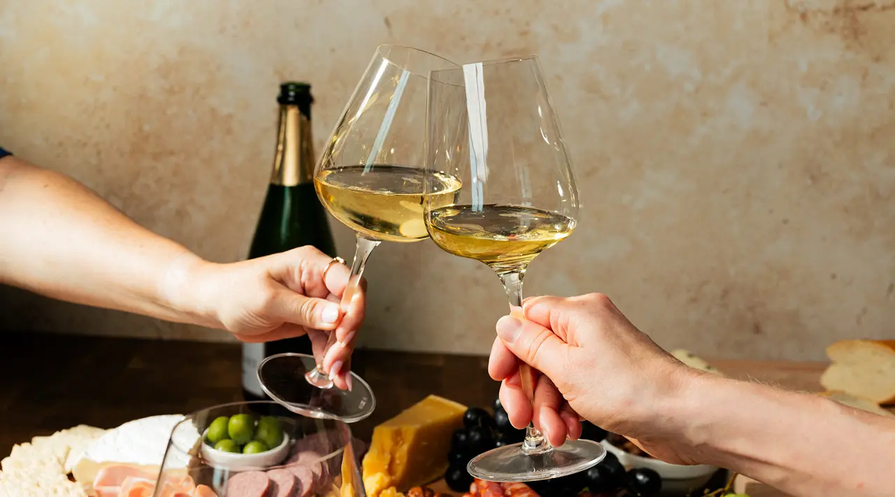 Two people are toasting with glasses of white wine over a table with a charcuterie platter in the background.