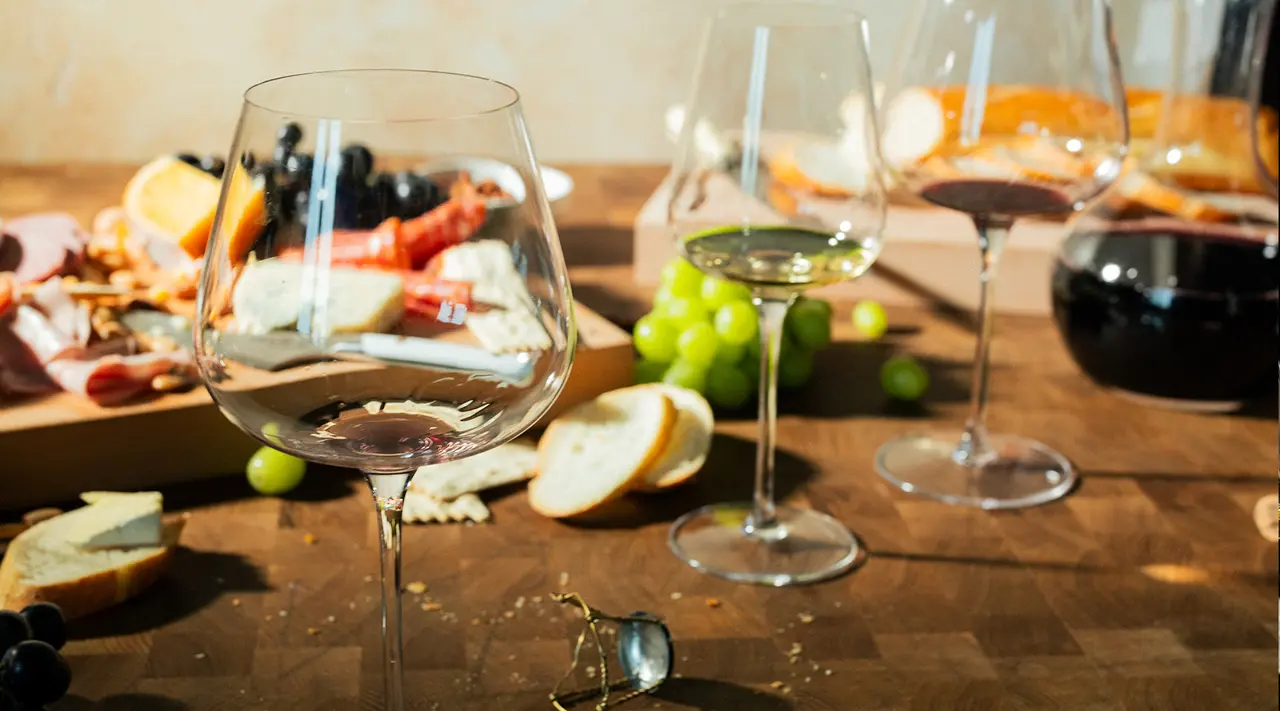 A spread of assorted cheeses, grapes, bread, and glasses of wine on a wooden table, evoking a sense of a gourmet tasting or social gathering.