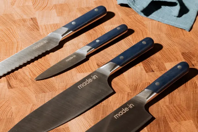 A collection of five kitchen knives with blue handles laid out on an orange textured surface, each stamped with the text "made in."