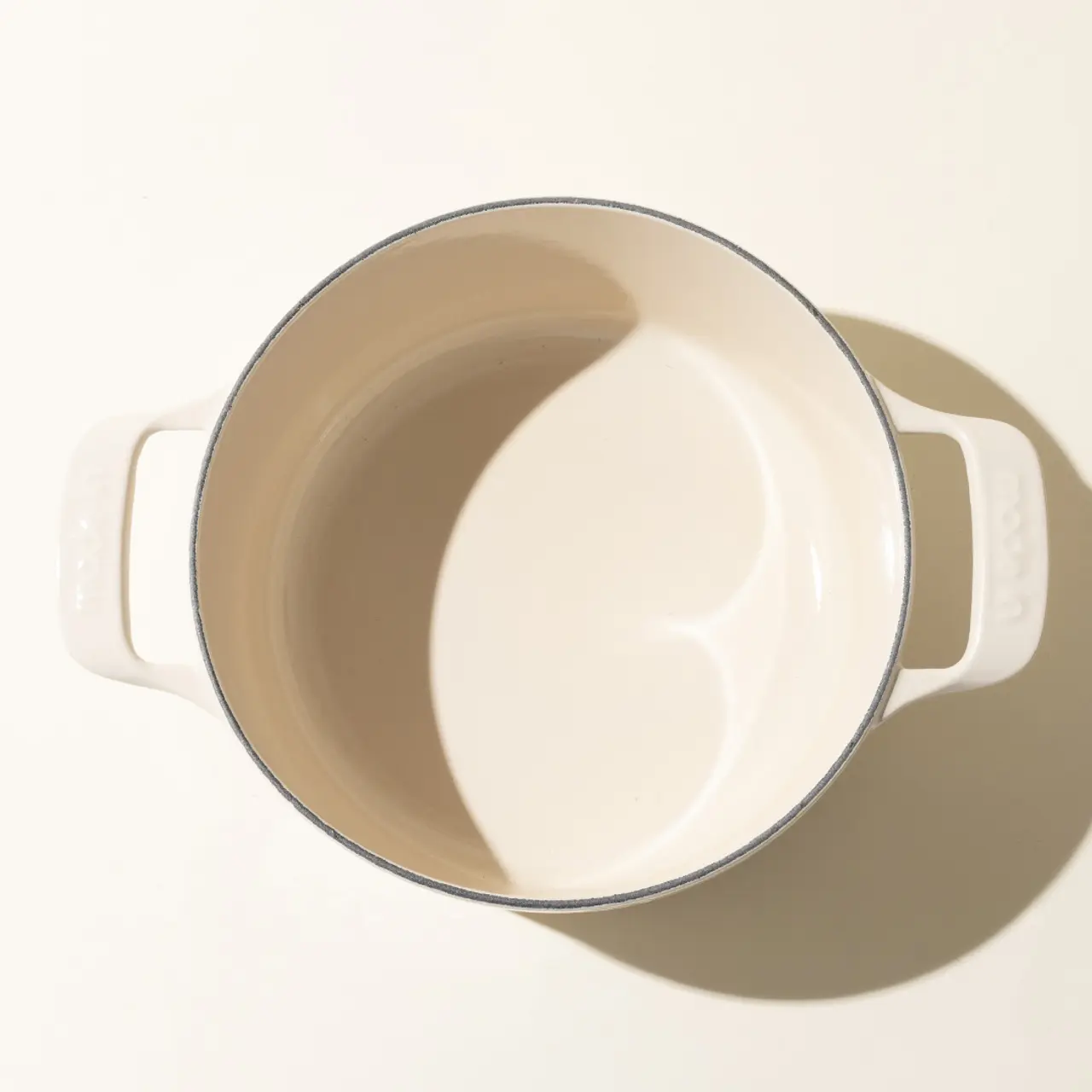 A cream-colored enameled cast iron pot with two handles, set against a light background casting a soft shadow.