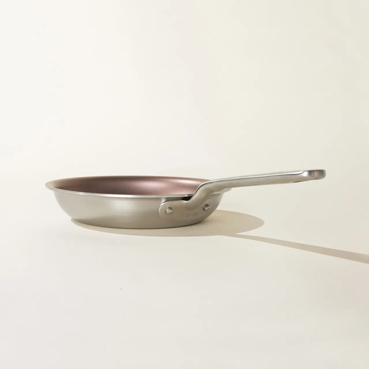 A stainless steel frying pan with a copper interior is placed against a neutral background, casting a soft shadow to one side.