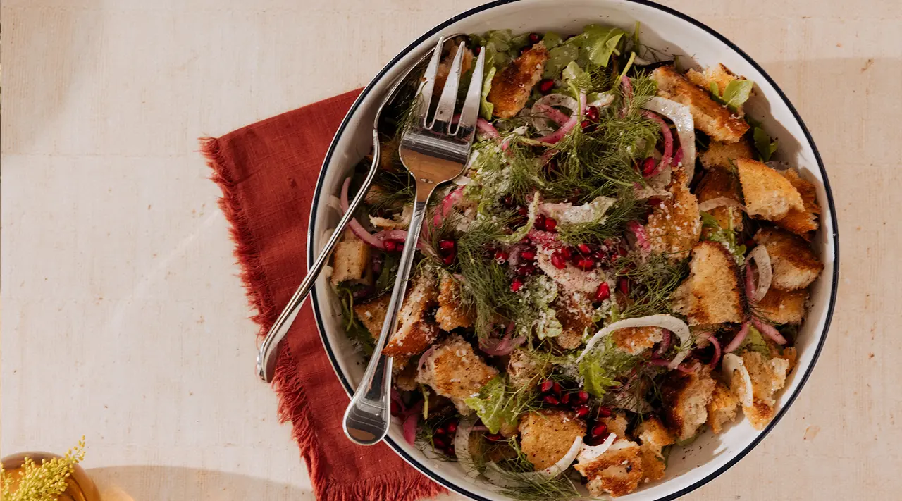 A bowl of leafy salad with croutons, slices of red onion, fresh herbs, and pomegranate seeds, served with a pair of salad servers on a tabletop with a terracotta cloth.