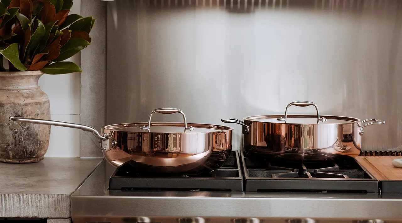 Two shiny copper pots sit on a modern stove next to a potted plant in a well-equipped kitchen.