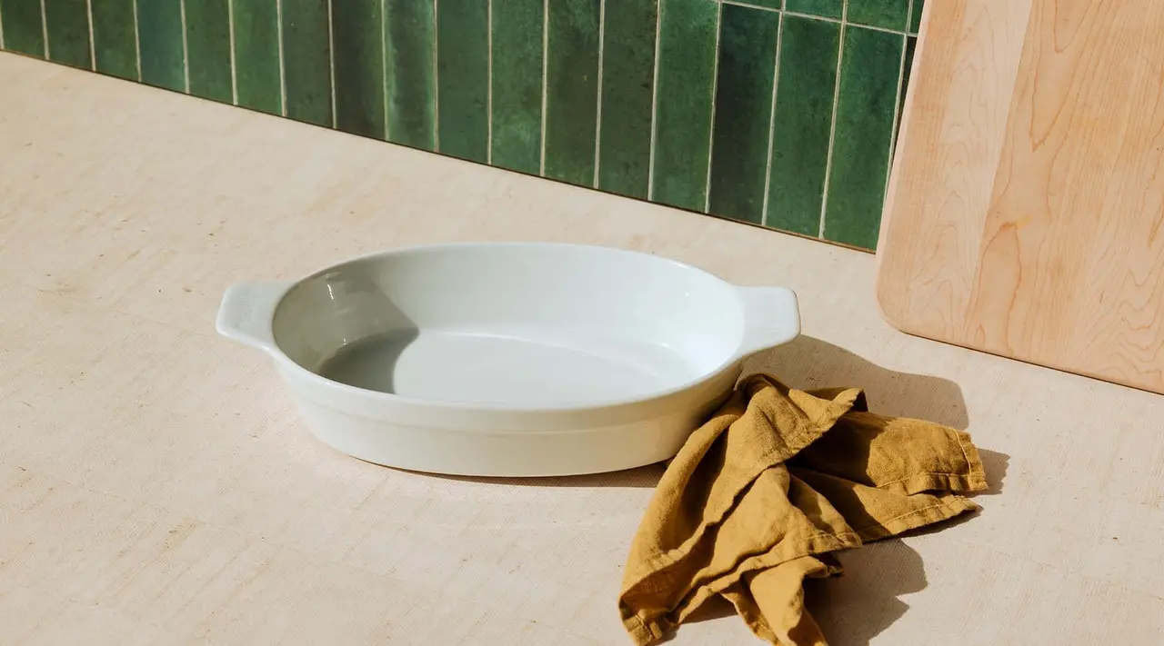 An empty white ceramic baking dish next to a crumpled brown cloth on a kitchen counter with green-tiled backsplash and a wooden board.