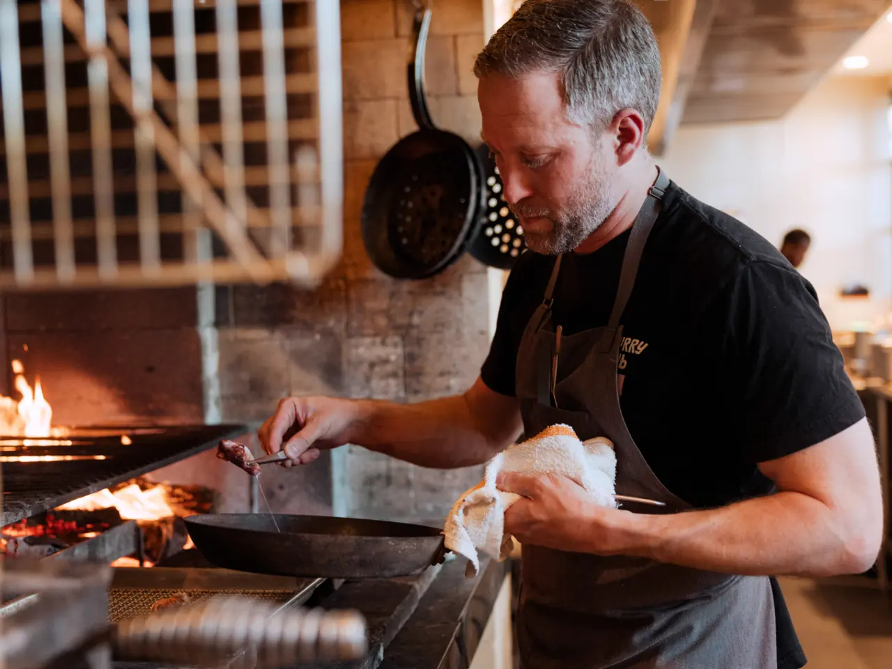 A focused chef attentively grills food over an open flame in a professional kitchen.