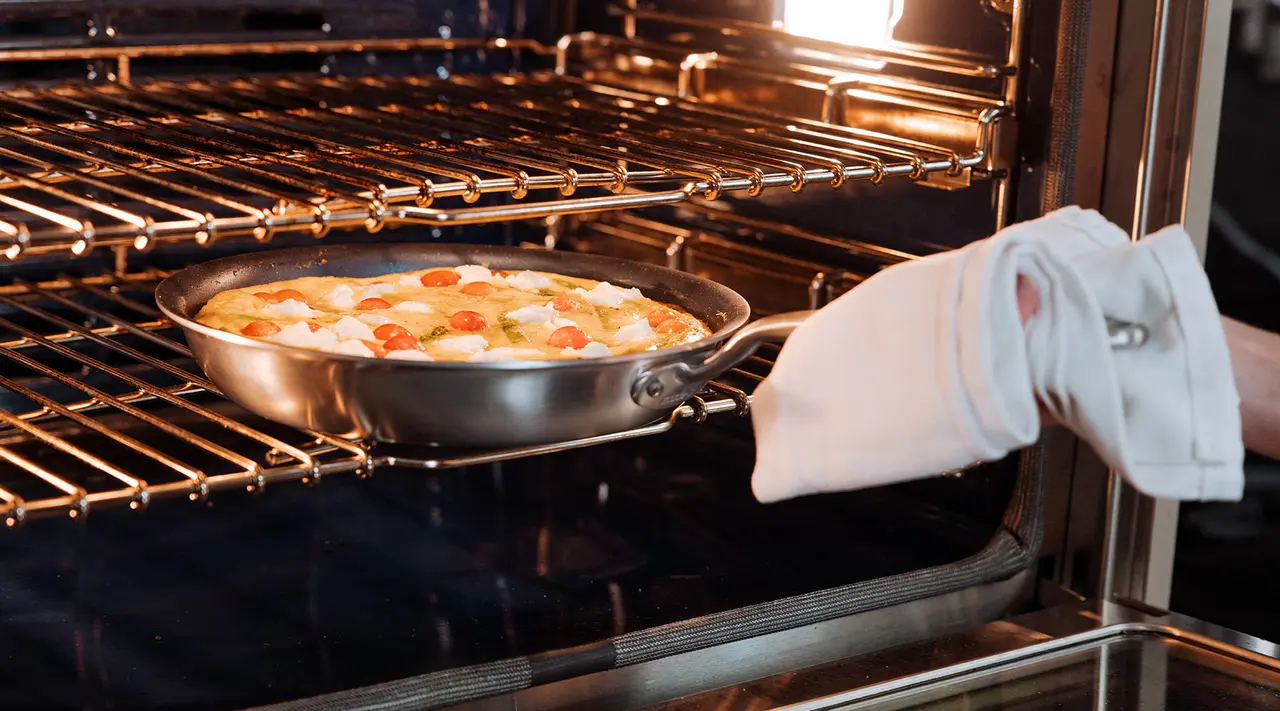 A hand wearing an oven mitt is sliding a skillet with a colorful frittata into a lit oven.