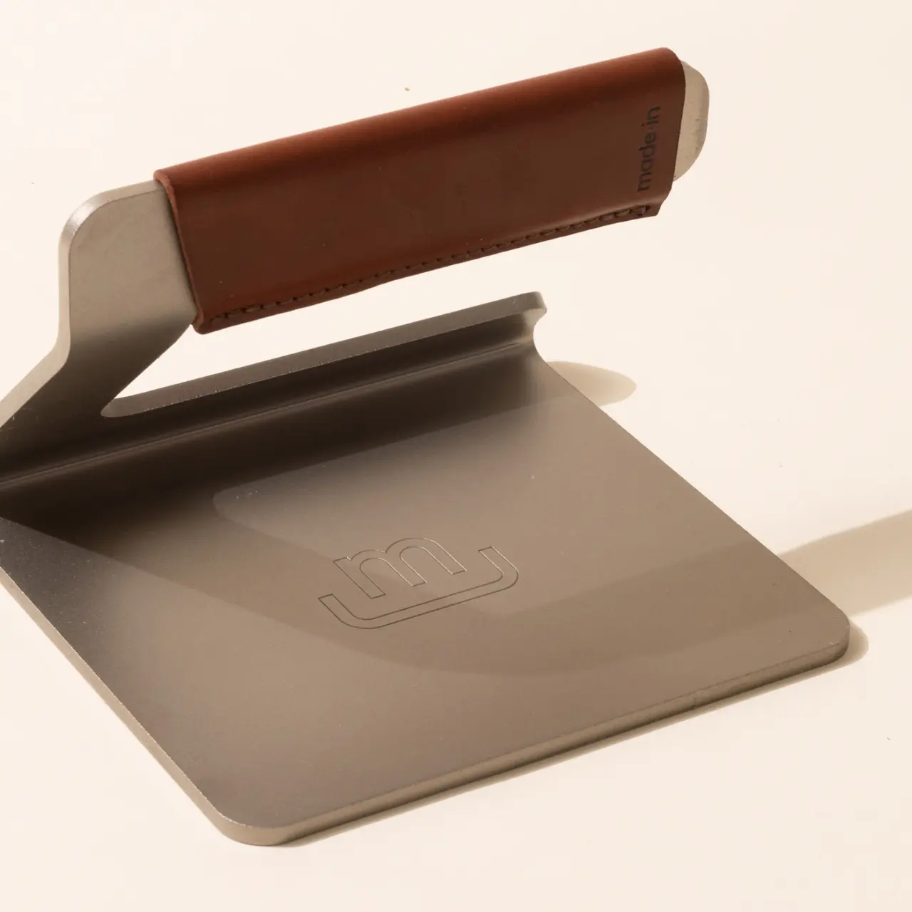A close-up of a sleek, modern hole punch with a metallic finish and a brown padded handle, casting a shadow on a plain surface.
