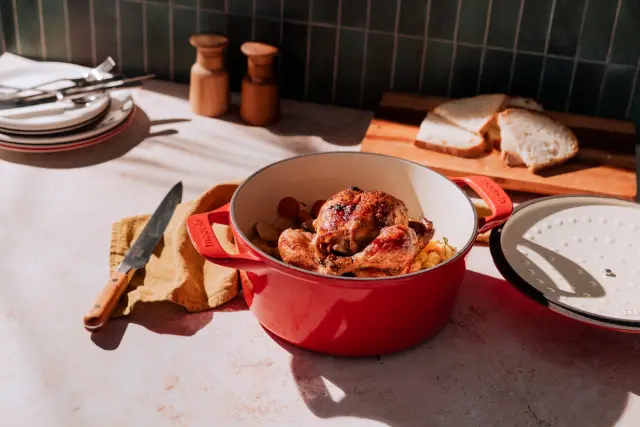 A freshly roasted chicken in a red pot on a sunlit kitchen counter with slices of bread and kitchenware.