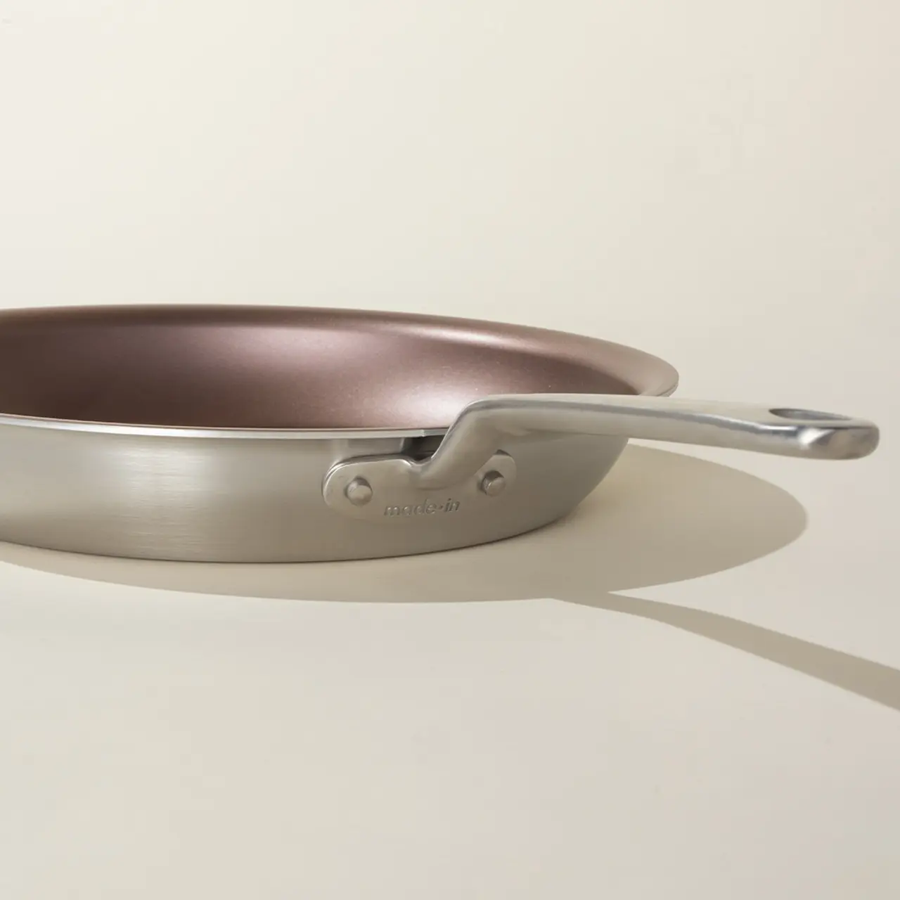 A stainless steel frying pan with a bronze-toned interior rests on a light surface casting a soft shadow.