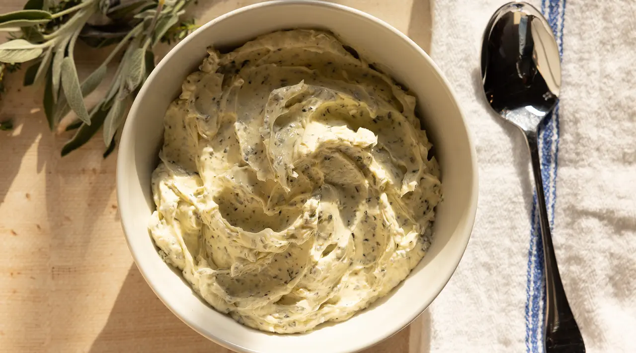 A bowl of creamy herb-infused dip with a spoon on the side, situated on a table with a cloth and some fresh herbs, under natural light.