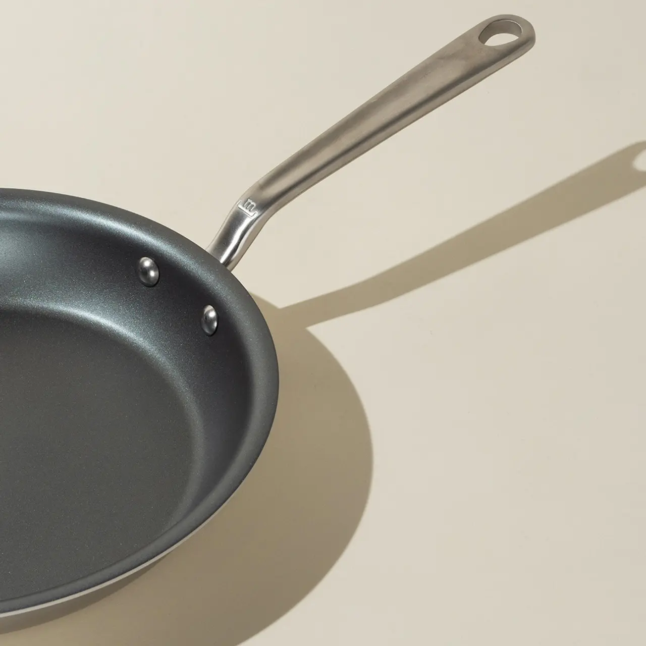 A non-stick frying pan with a stainless steel handle casts a shadow on a light surface.