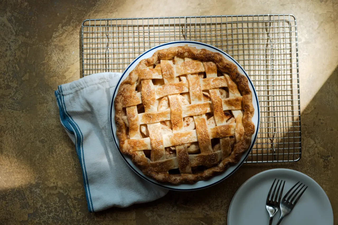 A freshly baked lattice-topped pie sits on a cooling rack next to a folded cloth napkin and a plate with a fork, all under warm lighting.