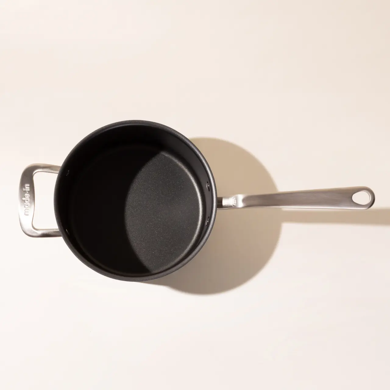 An overhead view of an empty saucepan with a long handle casting a shadow on a light surface.
