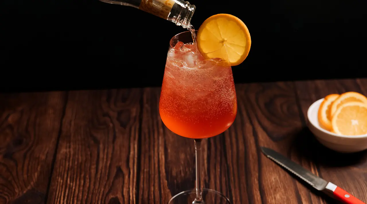 A refreshing cocktail with a slice of lemon is being poured into a wine glass on a dark wooden surface, with a bowl of sliced oranges and a knife to the side.