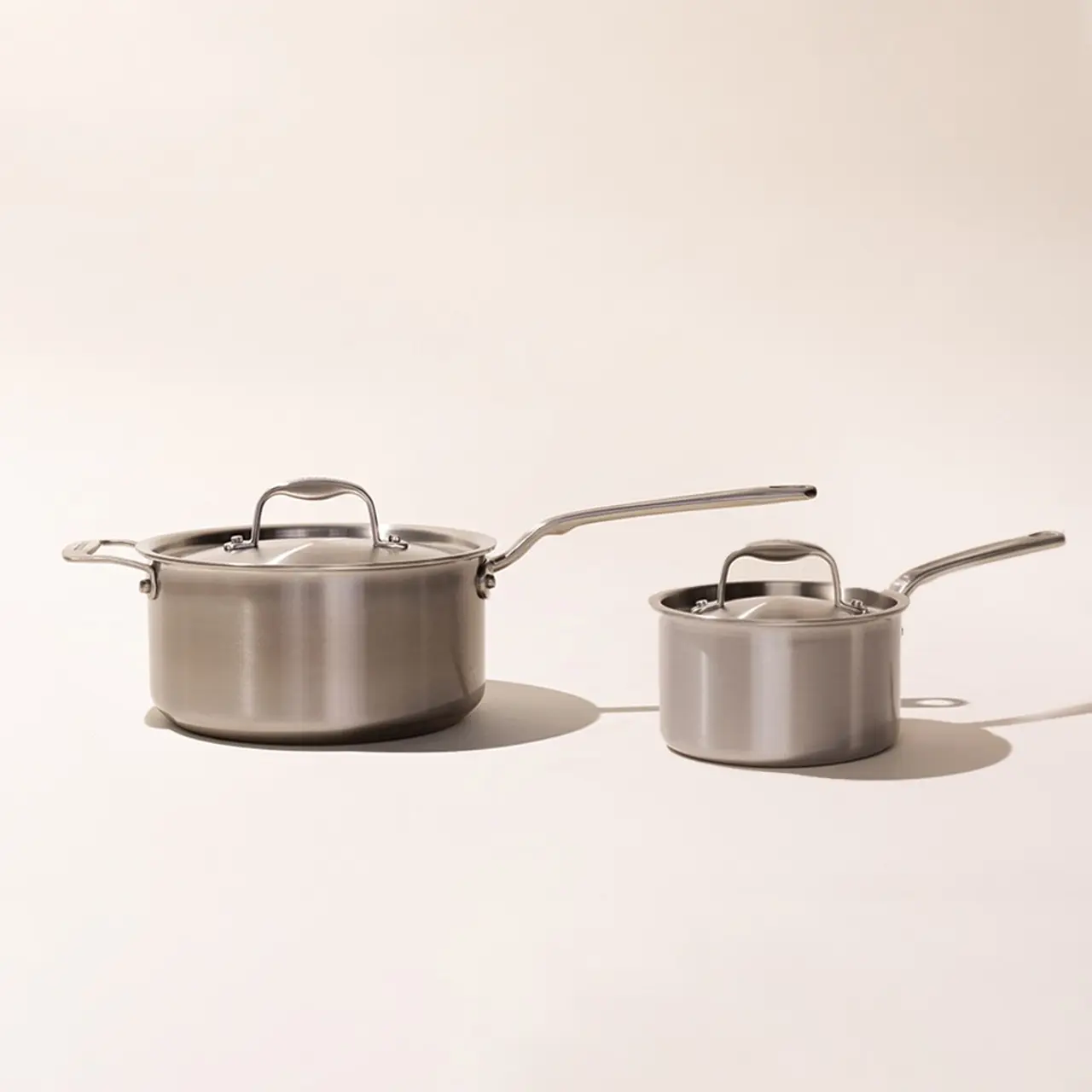 Two stainless steel pots with lids and long handles on a neutral background.