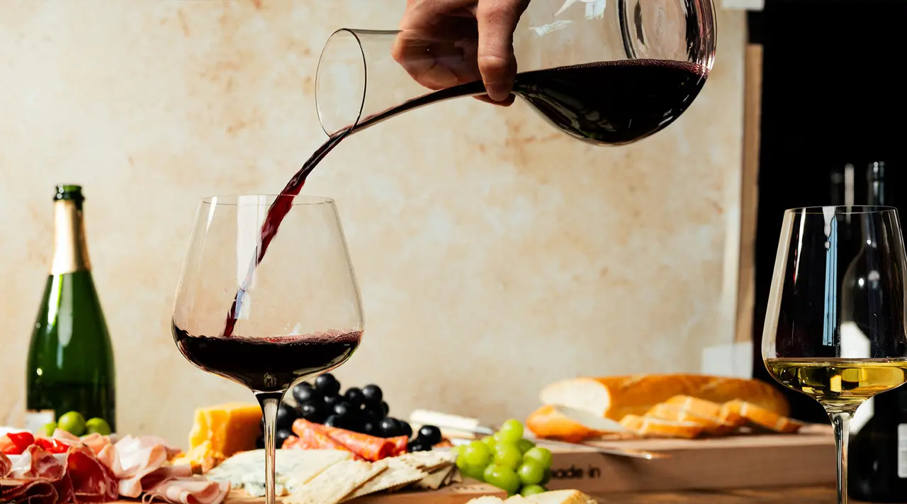 A person is pouring red wine into a glass with an assortment of cheeses, meats, bread, and grapes in the background.