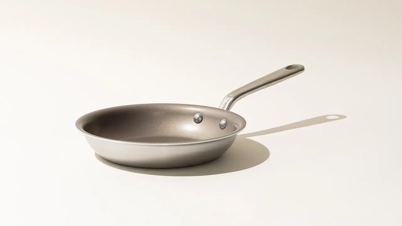 A stainless steel frying pan with a long handle casting a shadow on a light surface.