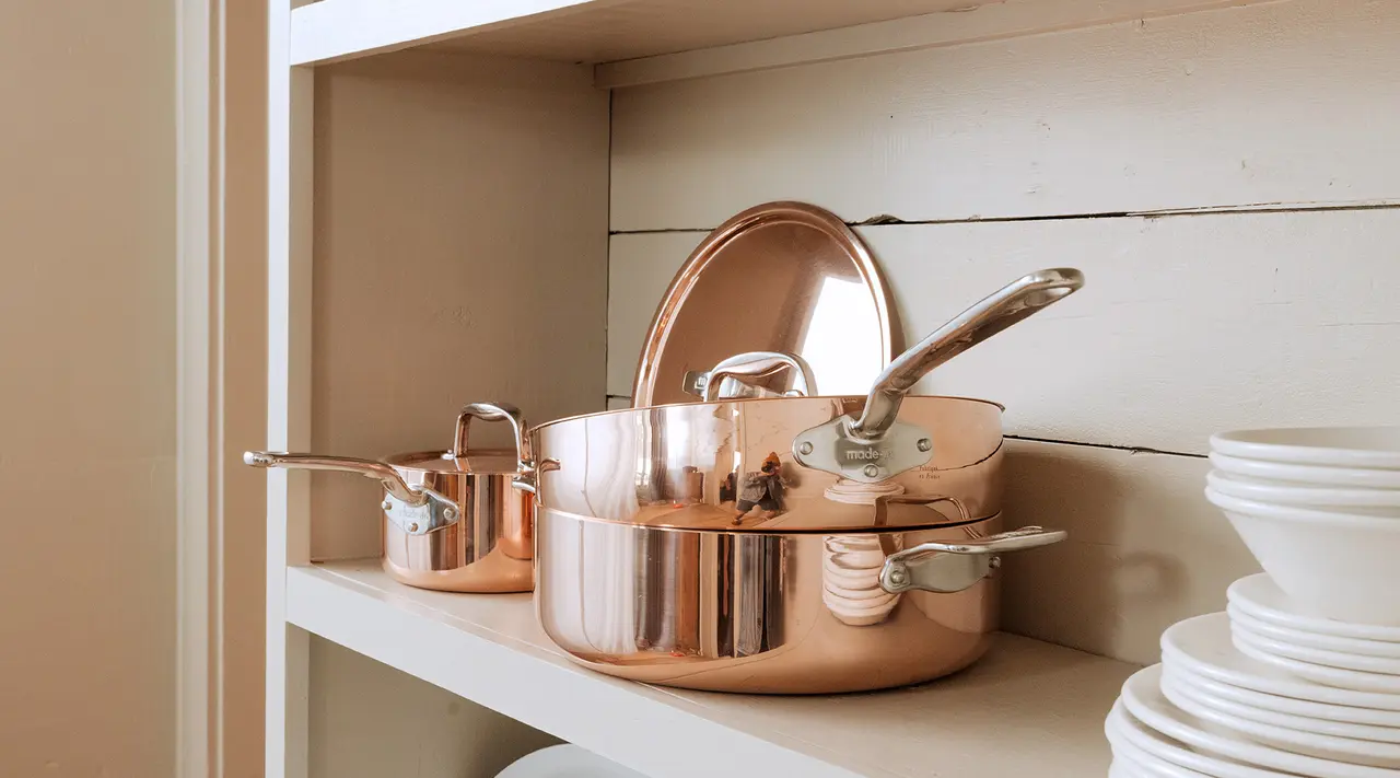 Shiny copper pots and pans are neatly arranged on a kitchen shelf next to a stack of white plates.