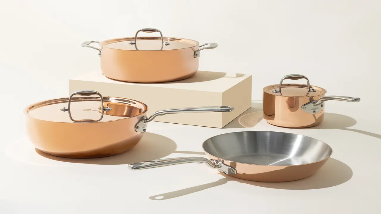 A set of four elegant copper-toned pans with stainless steel handles, including two pots with lids, a frying pan, and a small saucepan, displayed on a neutral background with a beige stand.