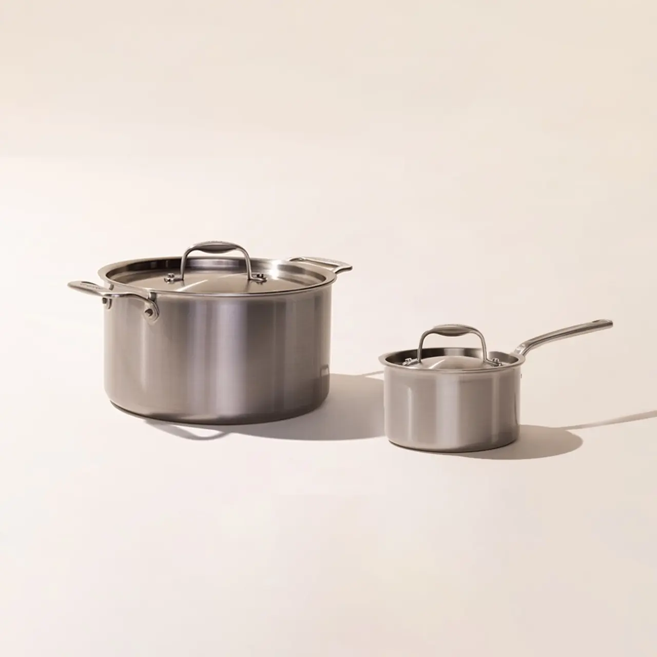 A large stainless steel pot with a lid and a smaller saucepan with a handle, both on a neutral background.