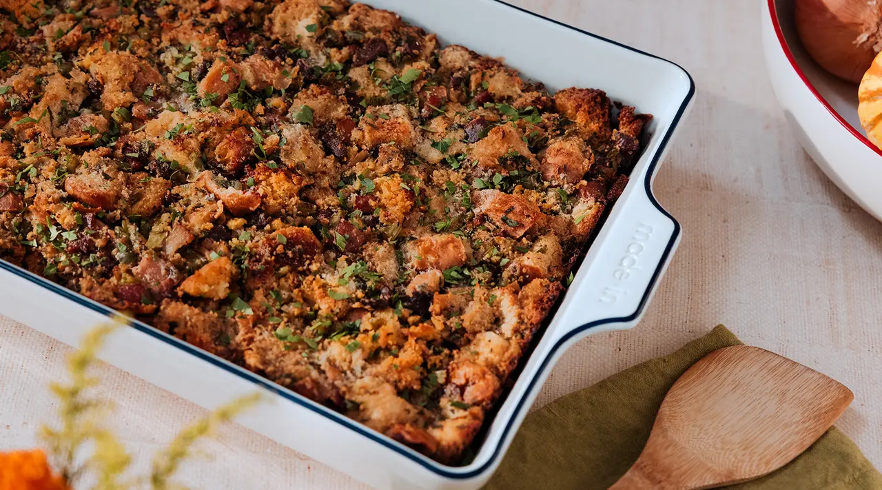 A freshly baked tray of stuffing garnished with herbs on a tablecloth, accompanied by a bowl and a wooden spoon.