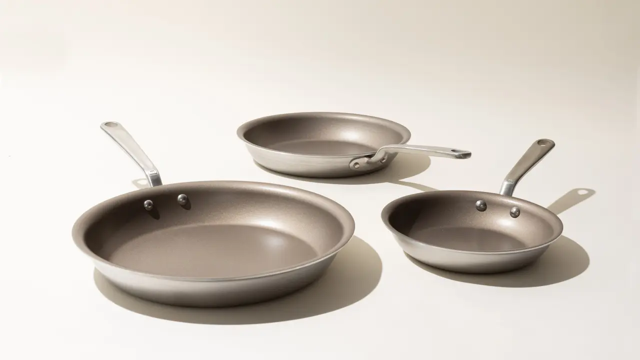 Three variously sized non-stick frying pans with stainless steel handles are arranged in descending order on a light surface against a white background.