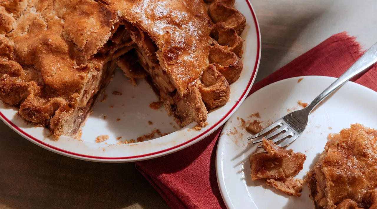 A freshly baked pie with a flaky crust is partially sliced on a plate next to a piece on a smaller plate with a fork.