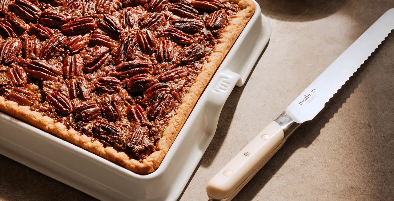 A freshly baked pecan pie in a white square baking dish next to a serrated knife on a kitchen counter.