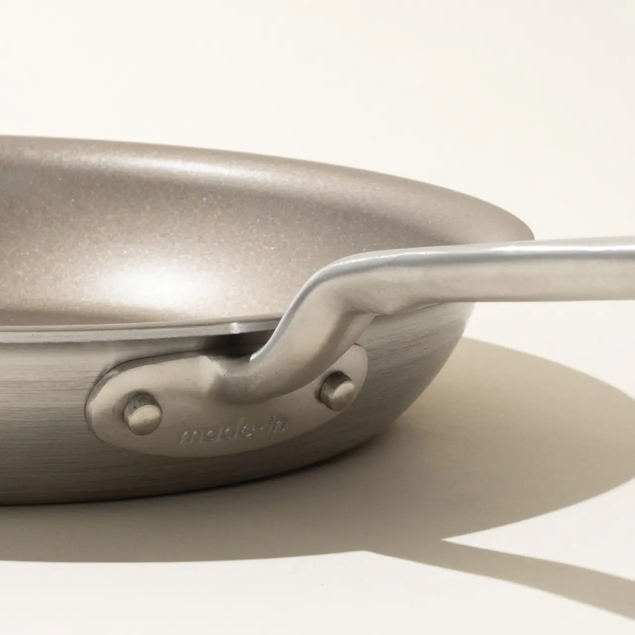 A close-up of a metallic frying pan focusing on its handle with the brand name visible and its shadow cast on the surface beneath it.