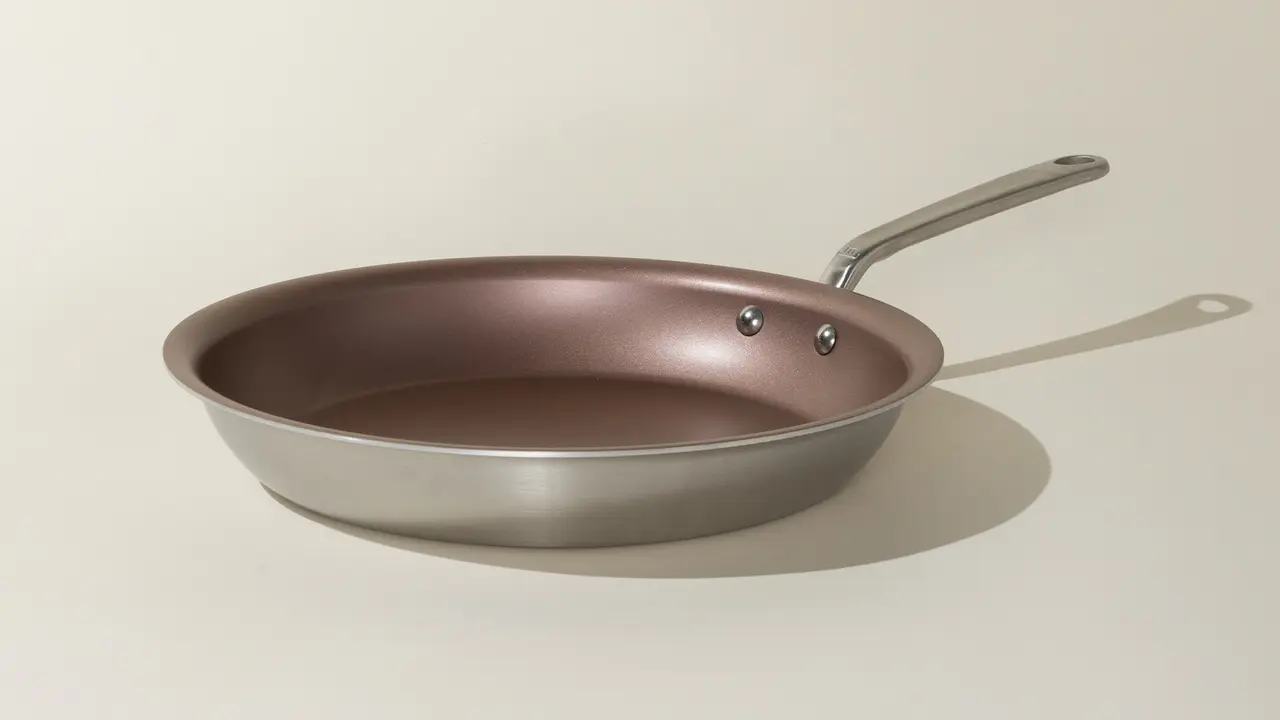 A silver and copper-toned frying pan with a long handle rests against a plain background.