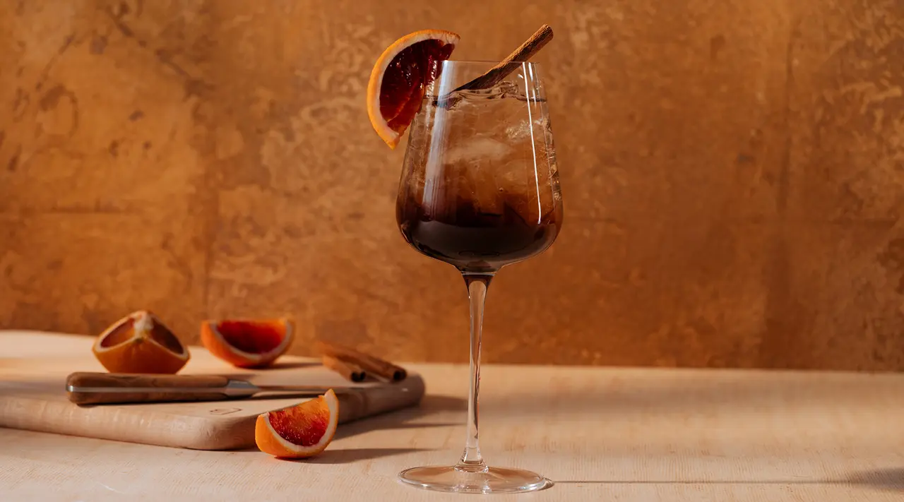 A wine glass filled with a dark liquid, garnished with an orange slice, against a warm textured background, with cut orange segments on a wooden board nearby.