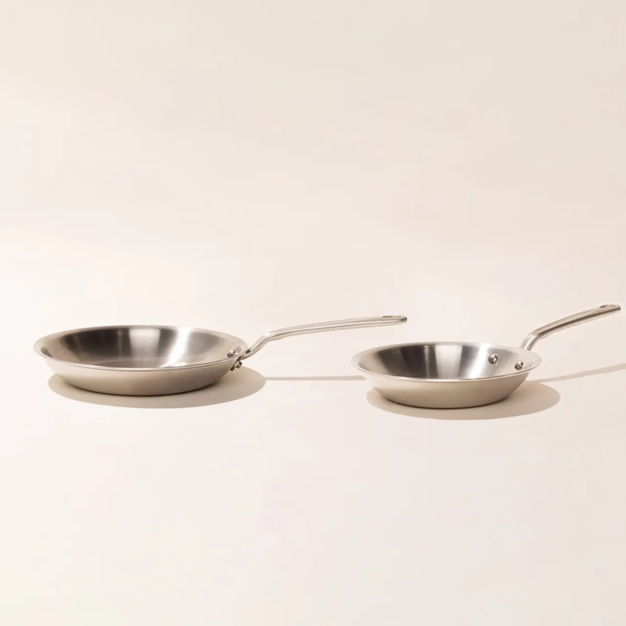 Two stainless steel frying pans with long handles are placed on a light-colored surface with a plain background.