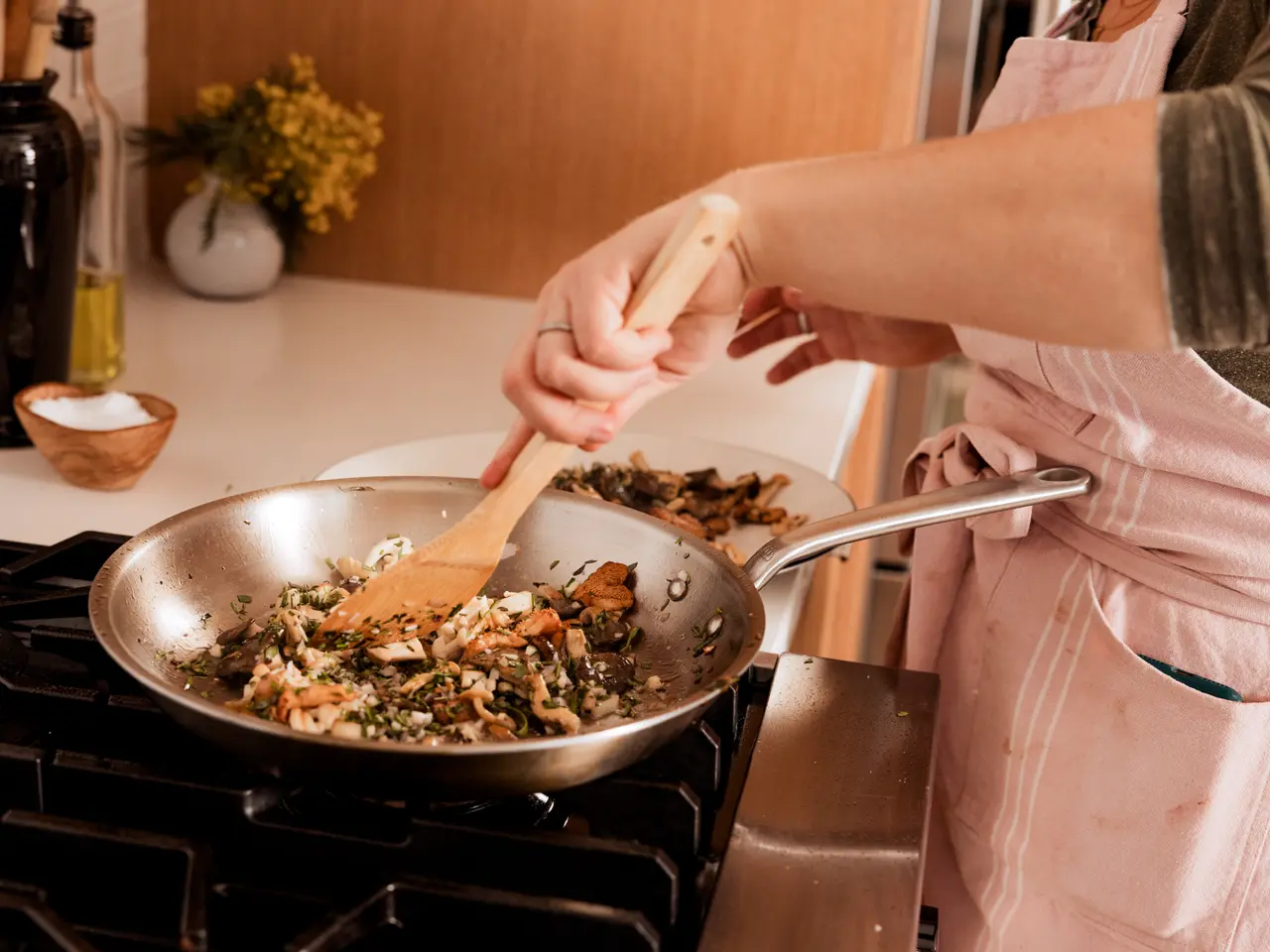 A person in a pink apron is sautéing food in a stainless steel frying pan on a stove.