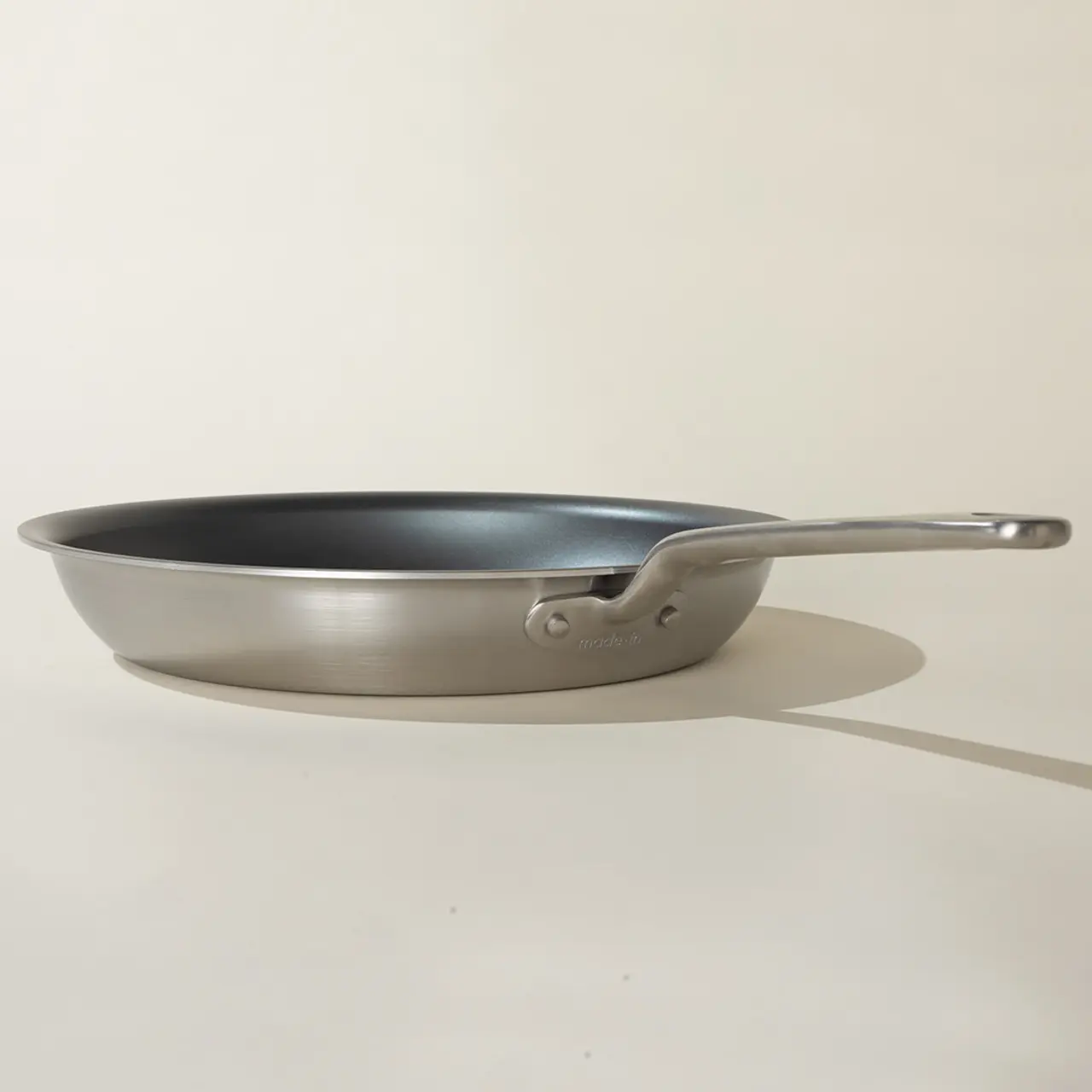 A silver frying pan with a long handle is positioned on a light background, casting a soft shadow.
