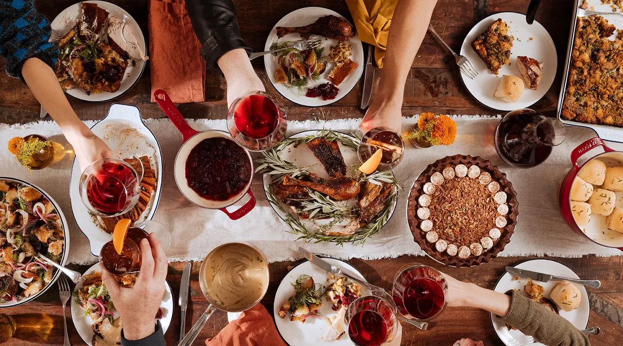 Overhead view of a festive table with people's hands serving and enjoying a variety of traditional Thanksgiving dishes.