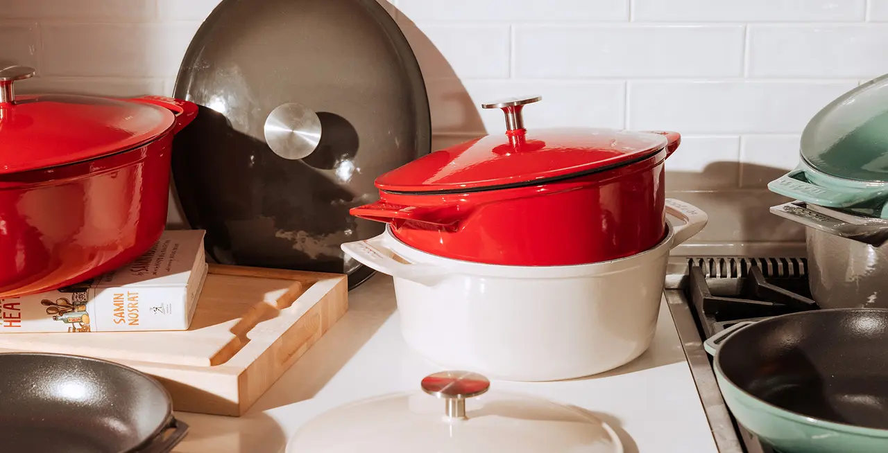 A variety of cookware including red and white enameled pots, skillets, and a wok, arranged on a kitchen counter.