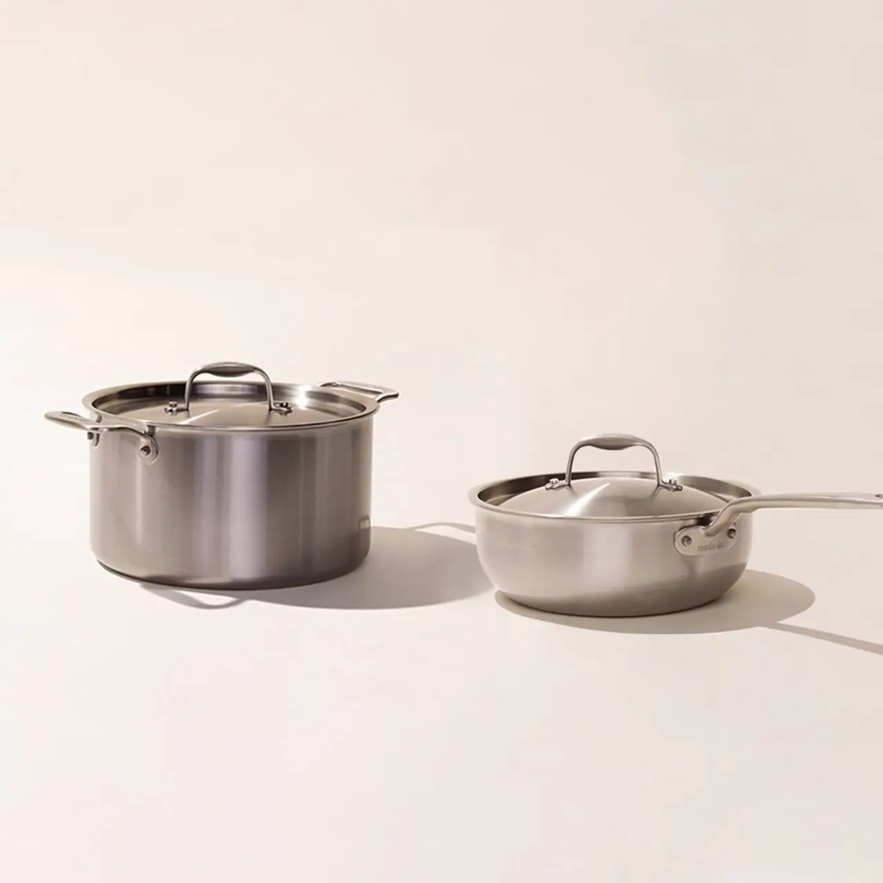 Two stainless steel cooking pots with lids, one with two handles and the other with a long handle, placed on a light background.