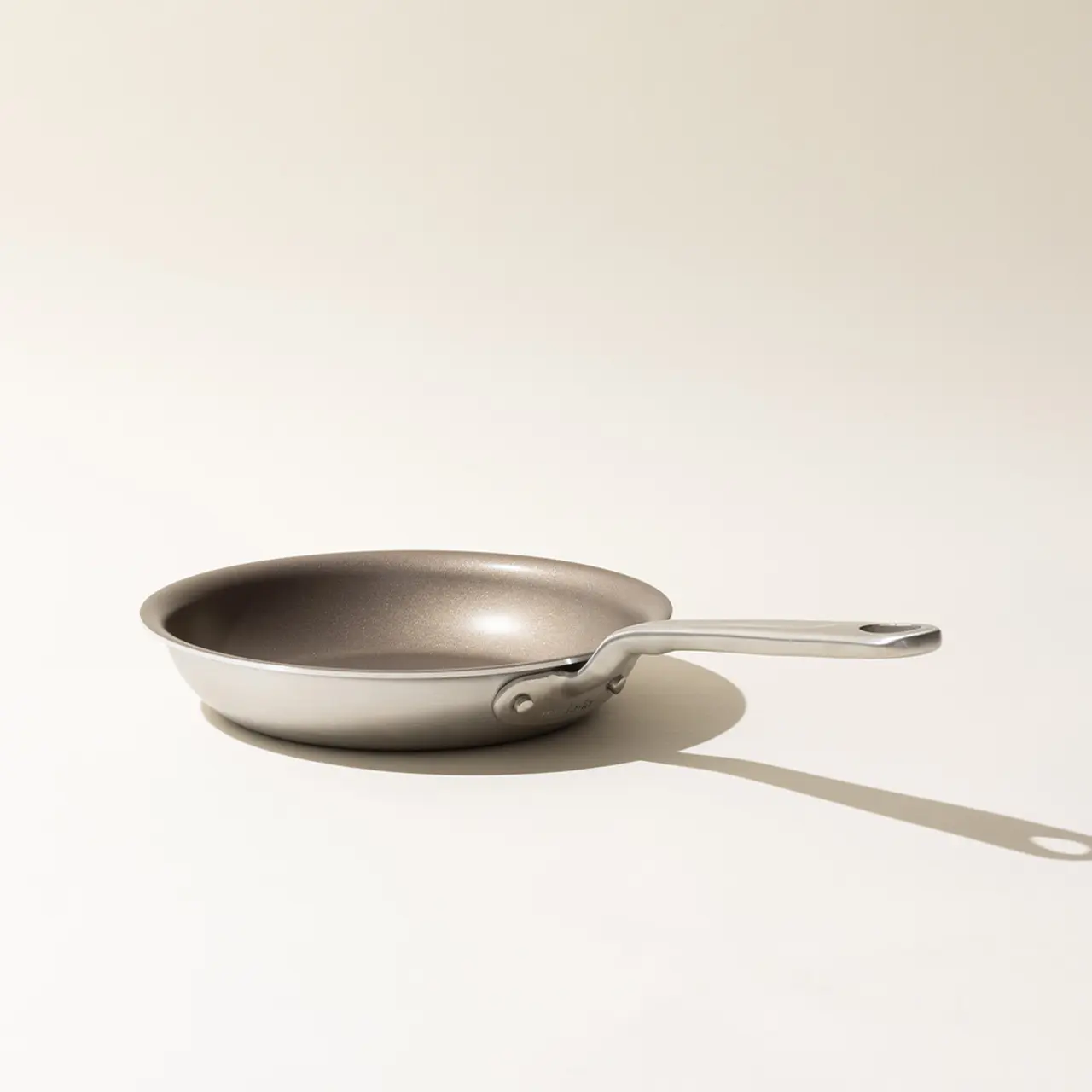 A stainless steel frying pan with a long handle is centered on a neutral background with a soft shadow indicating directional light.