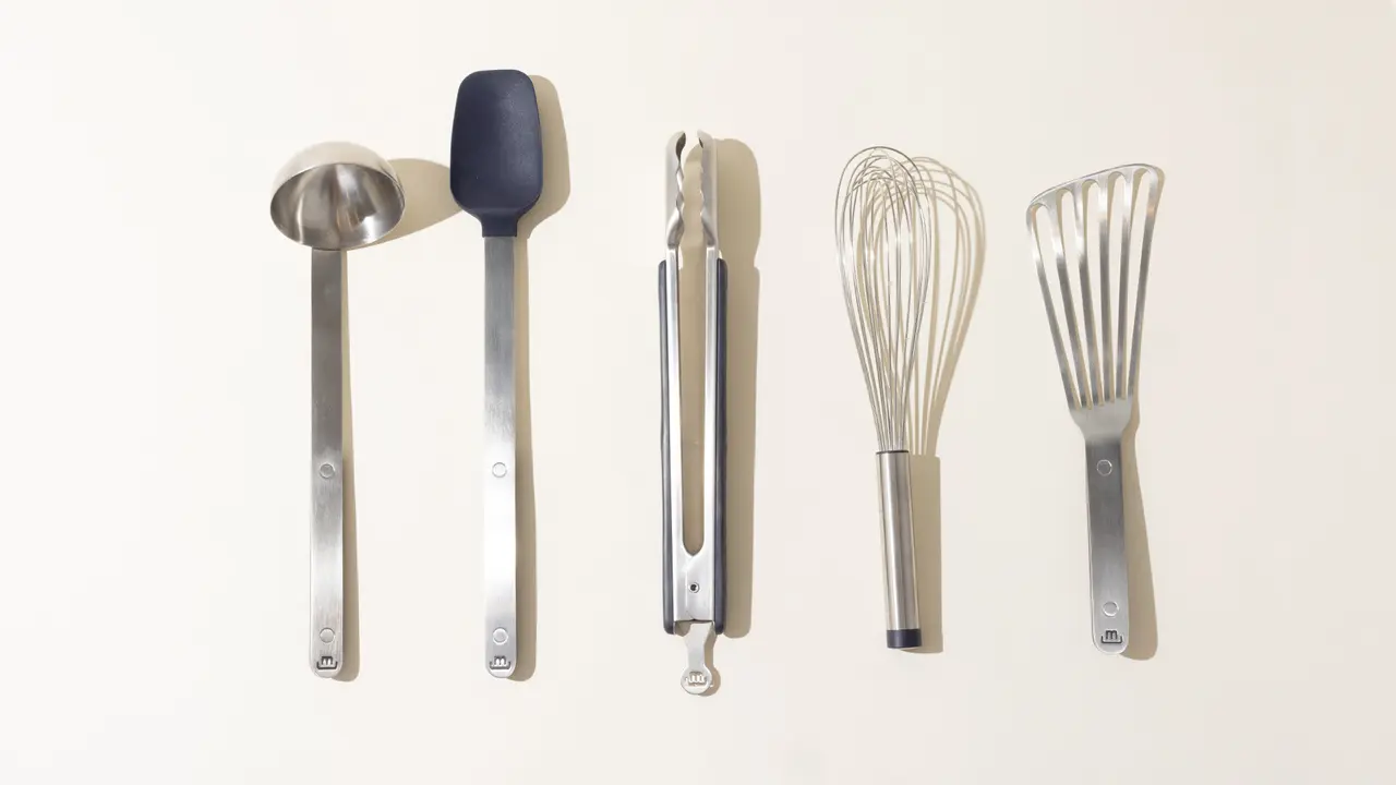 Five kitchen utensils, including a ladle, spatula, garlic press, whisk, and slotted spoon, are orderly arranged against a light background.