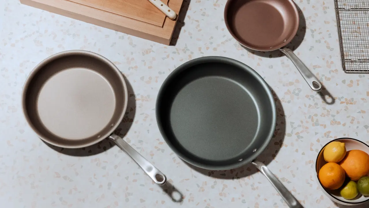 Three different-sized frying pans are laid out on a kitchen countertop next to a bowl of citrus fruits.