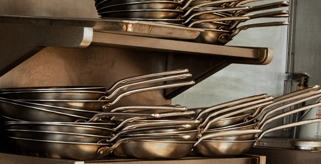 A stack of stainless steel frying pans on a shelf, with their handles aligned in a commercial kitchen setting.
