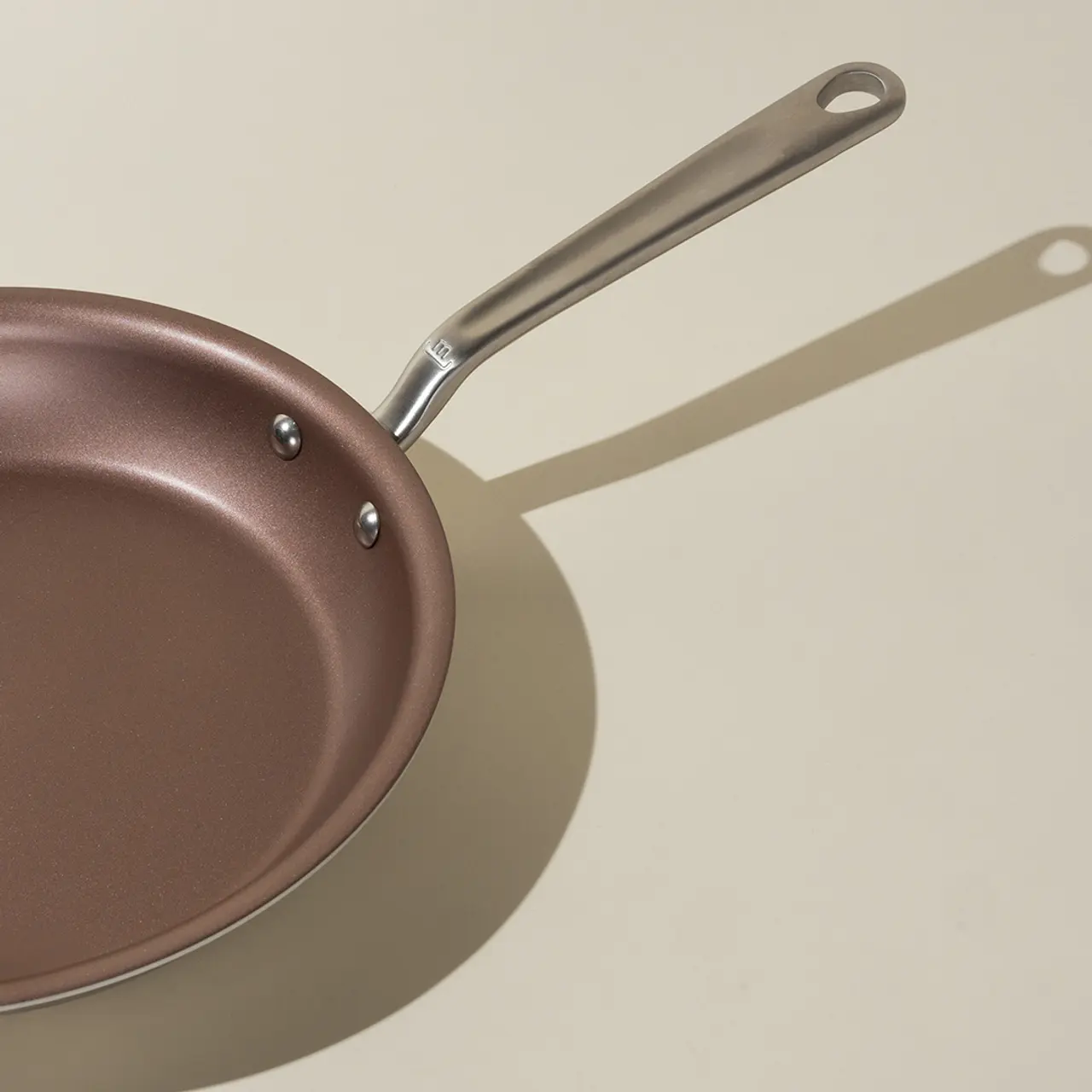 A non-stick frying pan with a stainless steel handle casts a shadow on a light surface.