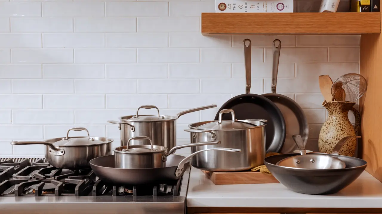 A variety of stainless steel and non-stick cookware neatly arranged on a modern kitchen stovetop and shelf against a white subway tile backsplash.