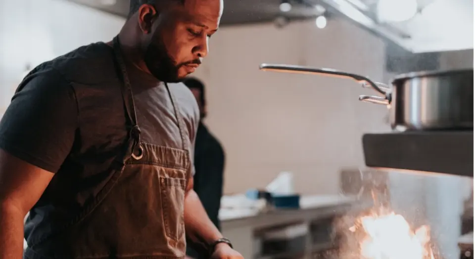 A focused chef wearing an apron observes a burst of flame from a pan while cooking.