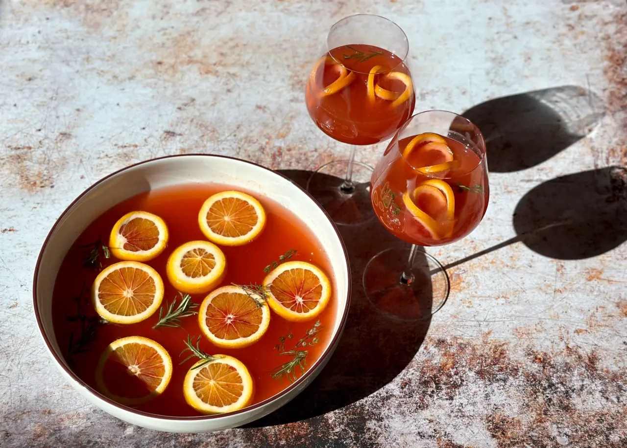 A punch bowl with orange slices and sprigs of rosemary is accompanied by two filled stem glasses on a textured surface, suggesting a refreshing beverage setup.
