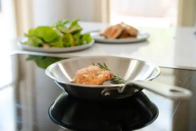 A piece of chicken with herbs is being cooked in a stainless-steel pan on a stove, with plates of salad and more chicken in the blurred background.